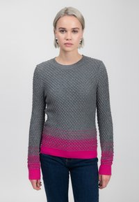 Vrouw met kort blond haar, die een gestructureerde grijze trui met een roze verloop aan de onderkant en manchetjes draagt, donkere jeans en zilveren oorbellen, staand tegen een witte achtergrond.