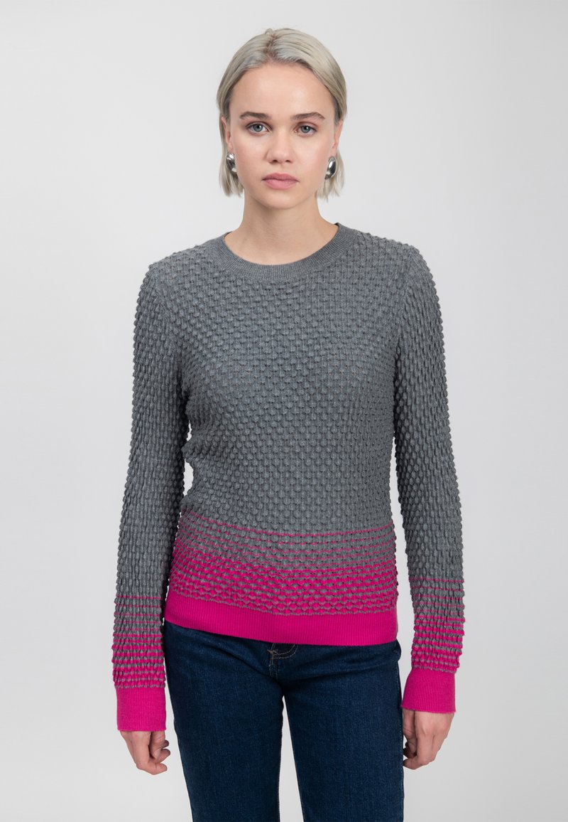 Vrouw met kort blond haar, die een gestructureerde grijze trui met een roze verloop aan de onderkant en manchetjes draagt, donkere jeans en zilveren oorbellen, staand tegen een witte achtergrond.