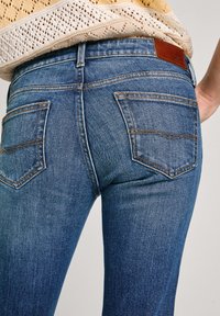 Primer plano de una persona que lleva unos pantalones vaqueros ajustados de denim azul con bolsillos traseros y una blusa de punto de ganchillo beige.