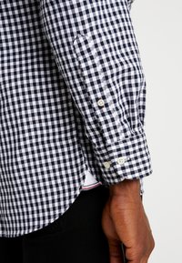 Chemise à carreaux bleu marine et blanche avec un poignet à bouton. Le tissu semble texturé avec un motif vichy classique. Fente latérale pour un meilleur ajustement.