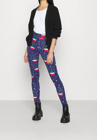 Leggings in dark blue with snow globe patterns featuring green trees in pots, complemented by a black oversized cardigan and chunky black boots.