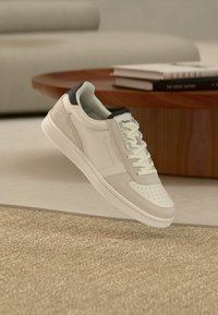 White leather sneaker with grey suede accents and a navy heel tab. Perforated toe design, flat laces, and a textured white sole.