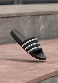 Sandale noire à glissière avec trois accents blancs rayés. Tige texturée et semelle plate pour le confort, présentant un design élégant et minimaliste.