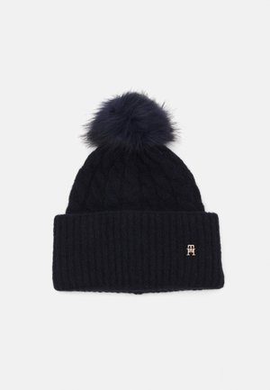 Beanie lavorato a maglia in blu navy con un bordo a coste e un motivo a treccia; presenta un grande pompon in pelliccia artificiale e una piccola accentuazione con logo dorato.