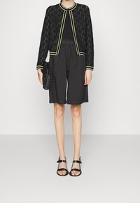 Czarna kurtka bomberka z żółtym wykończeniem, wzorzystym wzorem, w połączeniu z luźnymi czarnymi szortami. Czarne sandały na paskach i dopasowana torebka.