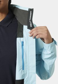 Světle modrá voděodolná bunda se zipovou boční kapsou, černými akcenty, měkkým límcem a viditelnou vnitřní etiketou.