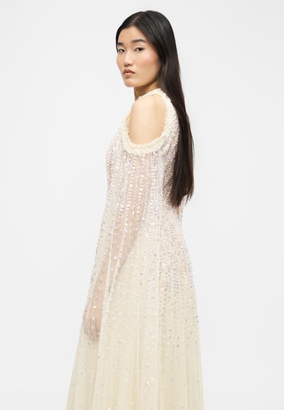 Giovane donna con lunghi capelli scuri che indossa un abito trasparente, con paillettes e spalle scoperte, vista di lato su uno sfondo semplice.