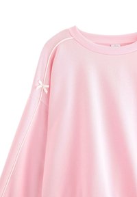 Rosa sweatshirt med ribbad rund hals, oversized passform och en dekorativ rosett på ärmen, med kontrasterande vit pipingdetalj.