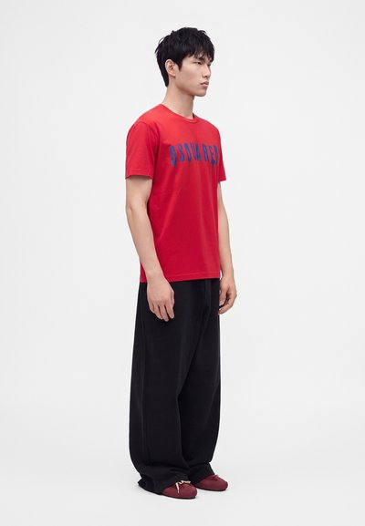 Un homme est debout de profil, portant un t-shirt rouge avec un texte bleu, un pantalon noir ample et des chaussures rouges sur un fond blanc uni.