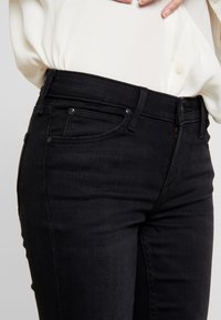 Jeans de mezclilla negra con un diseño ajustado, que presentan bolsillos frontales, cremallera y detalles de costura sutiles. El material es suave con una ligera textura.