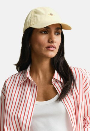 Frau trägt eine blassgelbe Mütze, weißes Tanktop und ein rotes und weißes, vertikal gestreiftes Hemd mit Knöpfen, schaut leicht zur Seite.