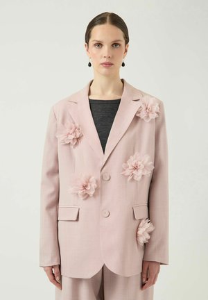 Femme portant un blazer rose clair avec de grandes fleurs en tissu cousues dessus, par-dessus un haut gris foncé, regardant droit devant avec une expression neutre.