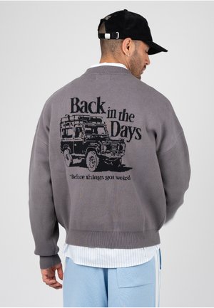Homme portant un pull gris avec un camion vintage noir et le texte "Back in the Days before things got weird,", casquette noire, chemise blanche et pantalon bleu clair.