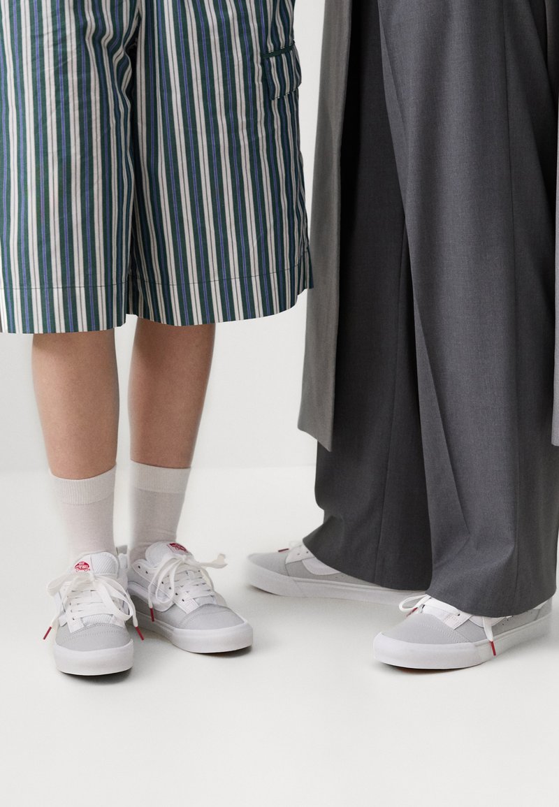 Zapatillas blancas y grises con acentos rojos, combinadas con pantalones cortos a rayas y pantalones grises. Incluye cordones y una suela texturizada.
