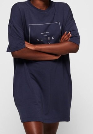 Person mit verschränkten Armen, trägt ein locker sitzendes, marineblaues T-Shirt-Kleid mit Text in einem Quadrat auf der Brust.