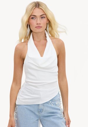 Femme blonde portant un top dos nu blanc et un jean bleu clair décoré de perles sur les côtés.