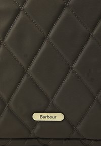 Tűzött sötétbarna anyag, gyémántmintázattal, középen egy kis arany fém Barbour logó táblával.
