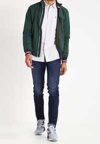 Muž v zelené bomber bundě přes bílou košili na knoflíky, tmavě modré džíny a šedé tenisky stojí před jednobarevným pozadím.