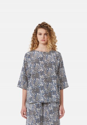 Donna con capelli biondi e ricci che indossa un top a fiori blu e bianco e pantaloni a gamba larga abbinati su uno sfondo uniforme.