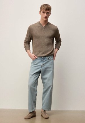 Giovane uomo in piedi con le mani nelle tasche, indossa un maglione marrone a scollo a V, jeans azzurri a vestibilità comoda e sneakers color cuoio, su sfondo neutro.