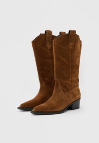 Ζευγάρι καφέ καουμπόικων μποτών από σουέντ με μυτερές μύτες, μεσαίου ύψους ενωμένες φτέρνες και θηλιές για να φοριούνται εύκολα, σε απλό φωτεινό γκρι φόντο.