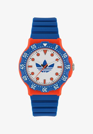 Analogni sat s bijelim licem, crvenim i plavim akcentima, plavim gumenim remenom i trokutastim markerom na 12 sati. Ima arapske brojeve.