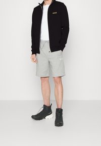 Svart zip-up jacka med guld "BOSS"-logotyp, vit t-shirt, grå fleece-shortsen och svarta robusta stövlar med vita sulor. Enkelt design, avslappnad passform.