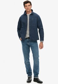 Marineblauwe jas met een zak aan de voorkant, knoopsluiting en kraag, gedragen over een grijze gebreide shirt. Combinatie met lichtblauwe jeans en zwarte laarzen.