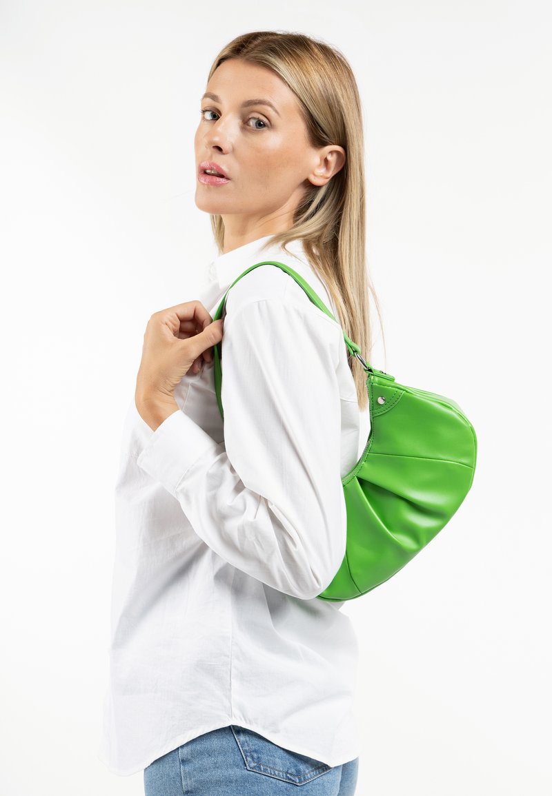 Grüne Leder-Hobo-Tasche mit gerafftem Design, glatter Textur und einem einzelnen Tragegurt. Das Modell trägt ein weißes Shirt und Blue Jeans.