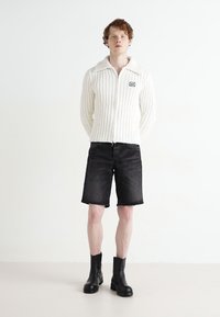 Suéter blanco de punto acanalado con cremallera frontal y cuello, combinado con pantalones cortos de mezclilla negros y botas negras hasta el tobillo.