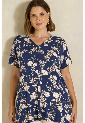 Femme portant un haut bleu marine à manches courtes avec un imprimé floral blanc et des boucles d'oreilles créoles dorées, debout devant un fond clair et neutre.