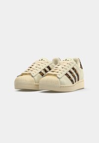 Beige leather sneakers with three brown stripes, textured rubber toe cap, and cream-colored sole. Laces and soft tongue complete the design.