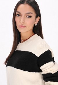 Stickad tröja i kräm- och svartrandigt mönster. Mjuk textur, rund halsringning och långa ärmar. Bärs med ett rött choker-halsband och silverfärgade hoopsörhängen.