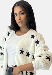 Vitt stickat cardigan med svarta stjärnmönster, ribbade mudd och en texturerad finish, lager ovanpå en figursydd vit topp.