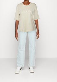 Beige kortærmet t-shirt med "CK" logo, iført lyseblå straight-leg jeans og hvide sneakers med sorte detaljer. Enkel, afslappet stil.