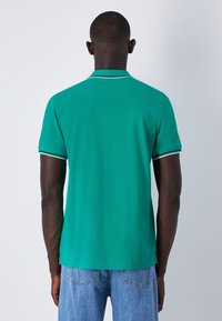 Grünes Polohemd mit einem Kragen und kurzen Ärmeln, ausgestattet mit kontrastierenden weißen Besätzen an den Ärmeln. Tragen mit hellblauen Jeans.