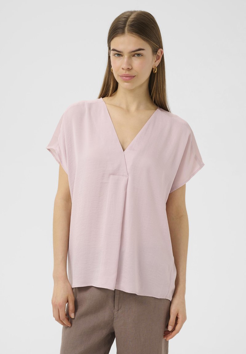 Blusa corta a maniche corte color rosa chiaro con scollo a V, vestibilità rilassata e taschino; realizzata in tessuto morbido e liscio.