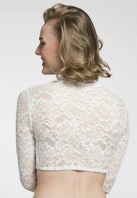 Weißes Spitzen-Crop-Top mit langen Ärmeln, das florale Muster und einen gewellten Saum aufweist. Es zeigt eine transparente Textur und eine figurbetonte Silhouette.