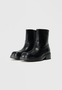 Botas negras hasta el tobillo hechas de cuero suave, con punta redondeada, cremallera lateral y un tacón bajo y grueso. Diseño minimalista con líneas limpias.