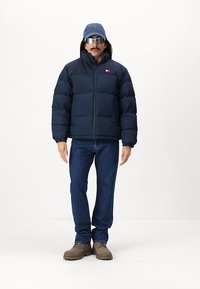 Marineblå puffjakke med høy krage, blå caps, solbriller, blå jeans og brune sko. Jakken har glidelås foran og logo-detalj.