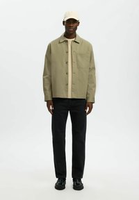 Homme portant une casquette beige, une veste boutonnée kaki clair sur une chemise crème, un jean noir et des chaussures noires, debout devant un fond blanc.