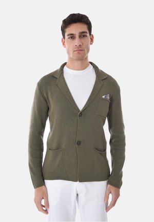 Giovane uomo che indossa un blazer a maglia verde oliva con due tasche e un fazzoletto nel taschino, abbinato a una maglietta bianca con scollo tondo e pantaloni bianchi.