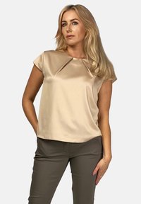 Beżowa satynowa bluzka z krótkimi rękawami, z unikalnym plisowanym wzorem przy dekolcie. Gładka tkanina, subtelny połysk i luźny krój.