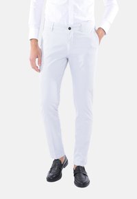 Pantaloni slim-fit di colore chiaro con una finitura in tessuto liscio, dotati di chiusura con bottone e tasche laterali, abbinati a scarpe nere senza lacci.