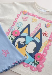 Rövid ujjú fehér póló kék rajzfilmes kutya grafikával, színes virágokkal és pillangókkal körülvéve, világoskék bordázott anyaggal párosítva.