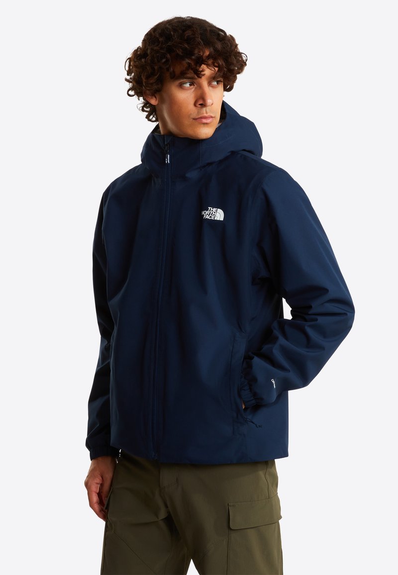 Veste imperméable bleu marine avec capuche, fermeture éclair, poignets élastiques et logo sur le côté gauche de la poitrine. Matière texturée, conçue pour un usage extérieur.