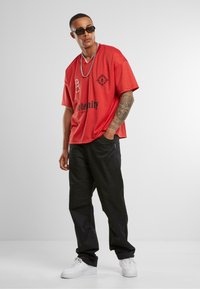Red oversized t-shirt with graphic designs and text, paired with black loose-fitting pants and white sneakers. Silver chain and sunglasses.