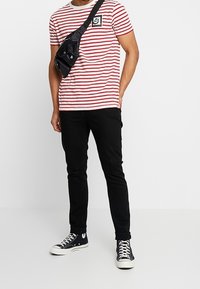 Červené a bílé pruhované tričko s kulatým výstřihem, kombinované s černými úzkými kalhotami a černými teniskami. Černá ledvinka je nošena přes tělo.