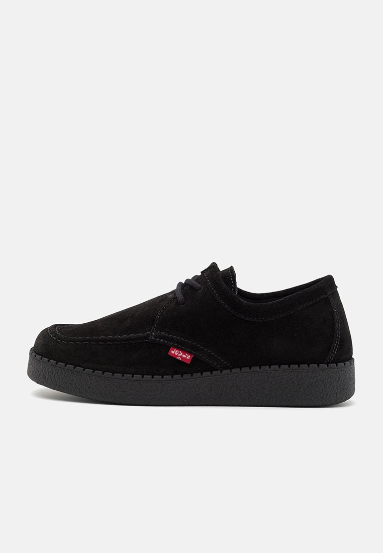 Chaussures en suede noir à lacets avec un bout arrondi, semelle texturée et une étiquette de logo rouge discrète sur le côté. Design profil bas.