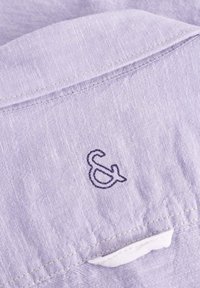 Tessuto viola chiaro con una sottile texture. Presenta un colletto e un piccolo logo ricamato in viola scuro "&" vicino al collo.